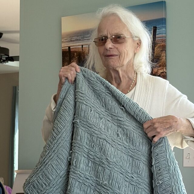 Gale Howell holding up a sage-green knitted sweater at a table.